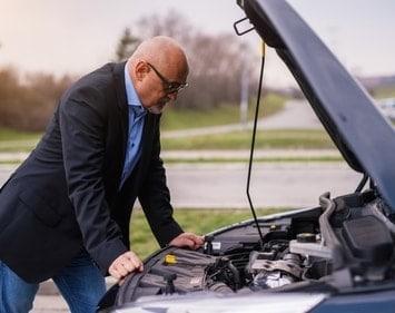 Man with glasses on the side of the road looking under his car